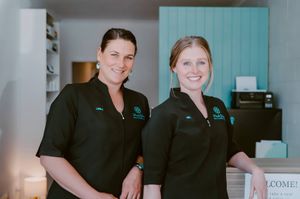 Two smiling women in matching black uniforms stand together at a welcoming reception desk in a modern salon.
