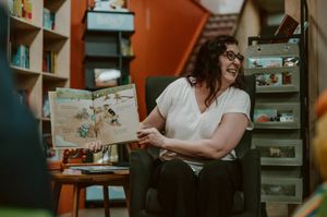 A woman laughing while reading a colorful picture book aloud in a cozy, book-filled room.