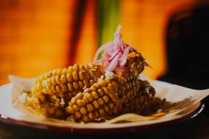 Grilled corn on the cob topped with pickled red onions and savory sauce served on a white plate.