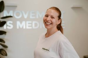 Smiling woman in white Bodyworx t-shirt standing in front of Movement Is Medicine wall sign.