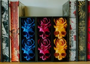 Three colorful skull and rose bookmarks in blue, pink, and gold displayed between gothic hardcover books on a shelf.
