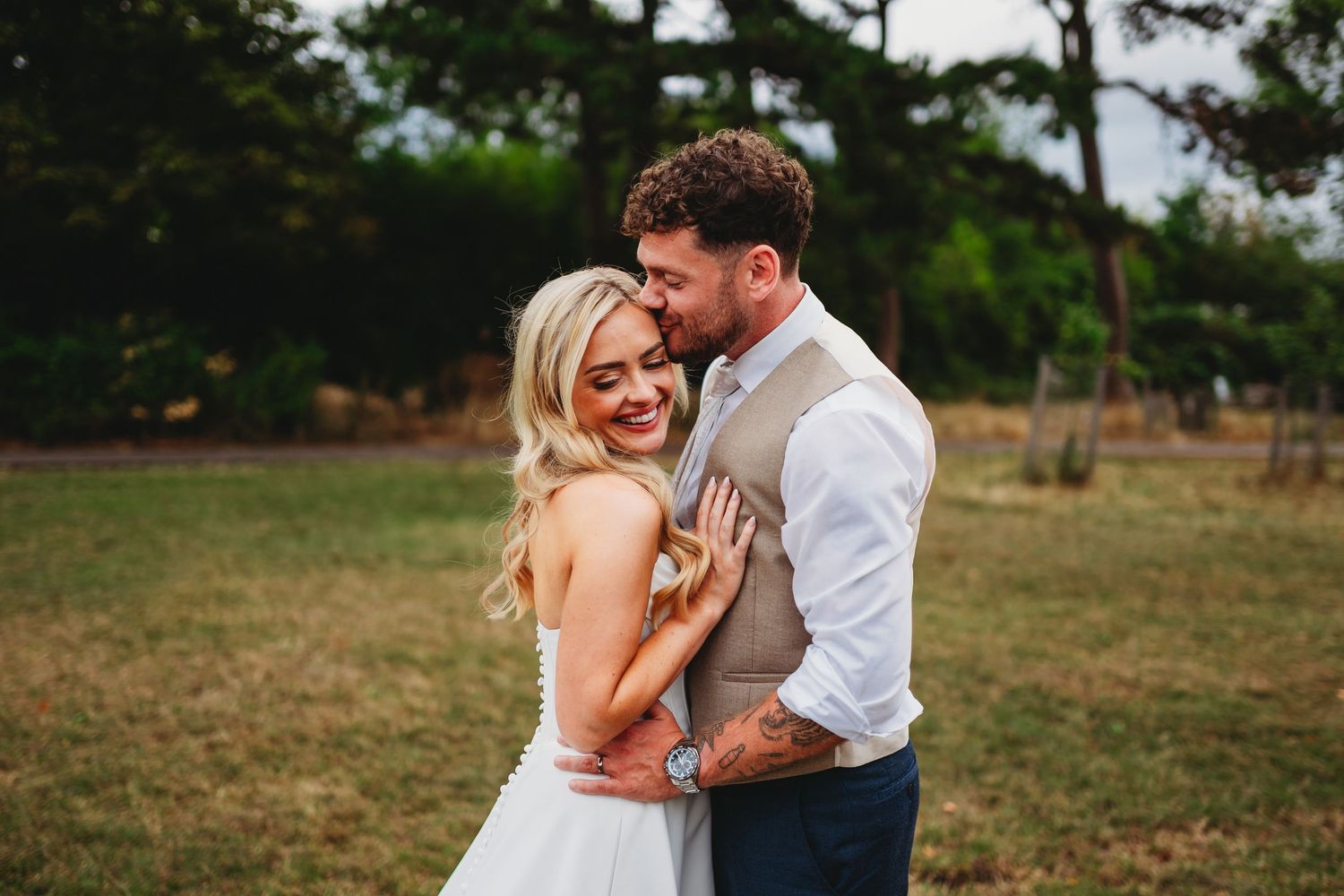 A groom in a vest embraces his smiling blonde bride in a white dress outdoors on their wedding day at Gloucester Old Spot