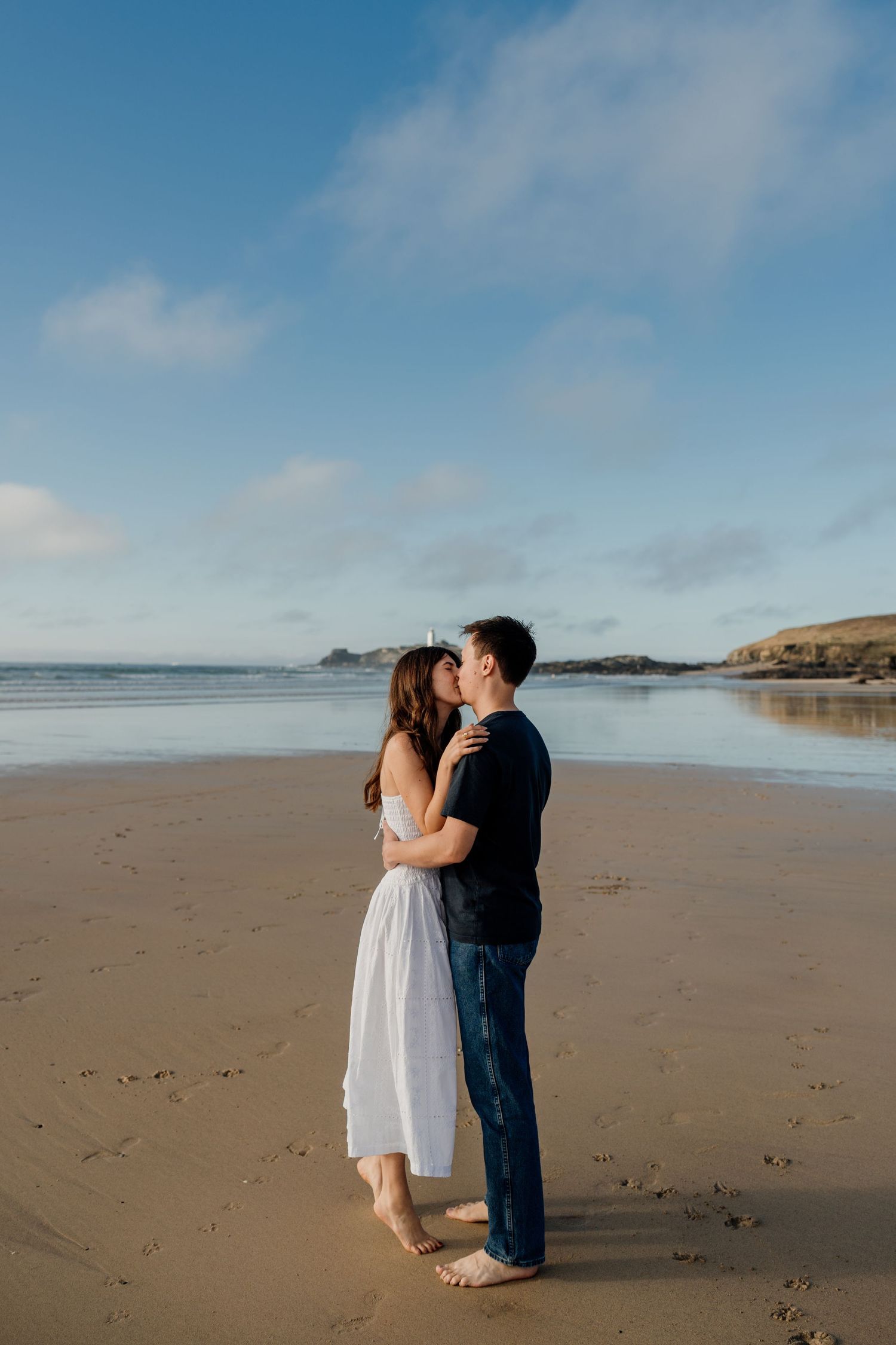 A couple shares a romantic kiss on a sandy beach with calm ocean waters and blue sky behind them.