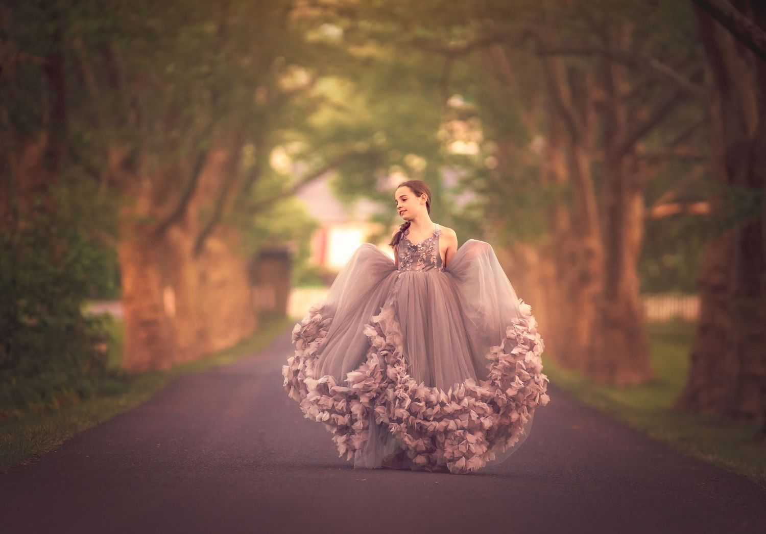 A woman in a flowing gray floral ball gown twirls on a tree-lined road in soft golden light.