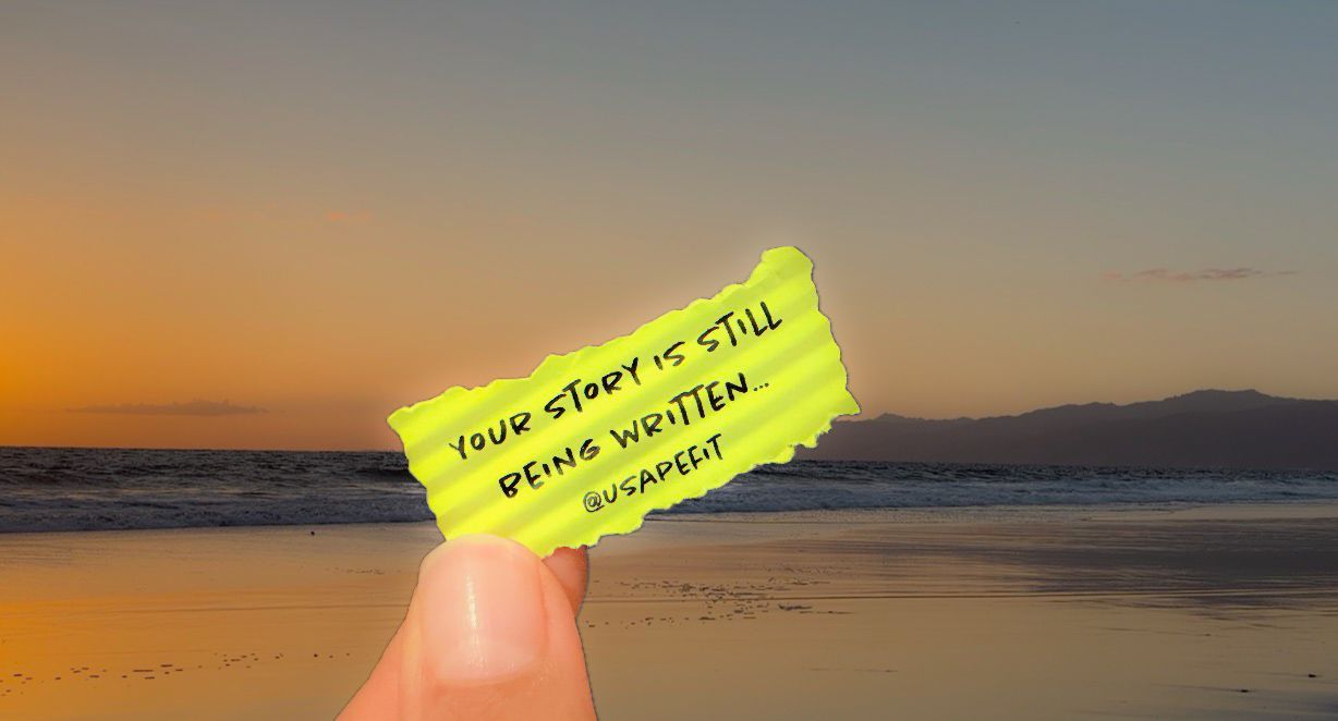 Hand holding a yellow note reading Your Story Is Still Being Written at a scenic beach sunset.