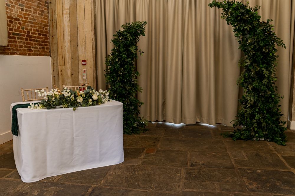Elegant wedding sweetheart table with white linen and floral centerpiece framed by lush greenery arch backdrop.