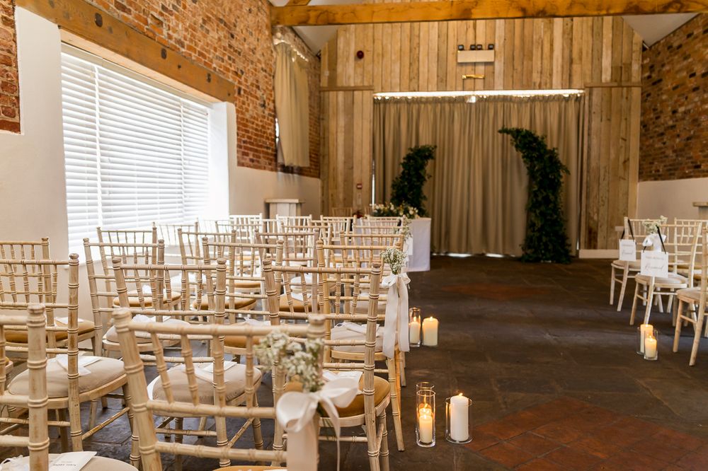 Elegant barn wedding ceremony setup with gold chiavari chairs, candles, and a floral arch backdrop.