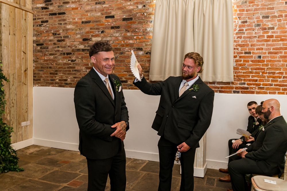 Two groomsmen in black suits share a lighthearted moment at a rustic brick-walled wedding venue.