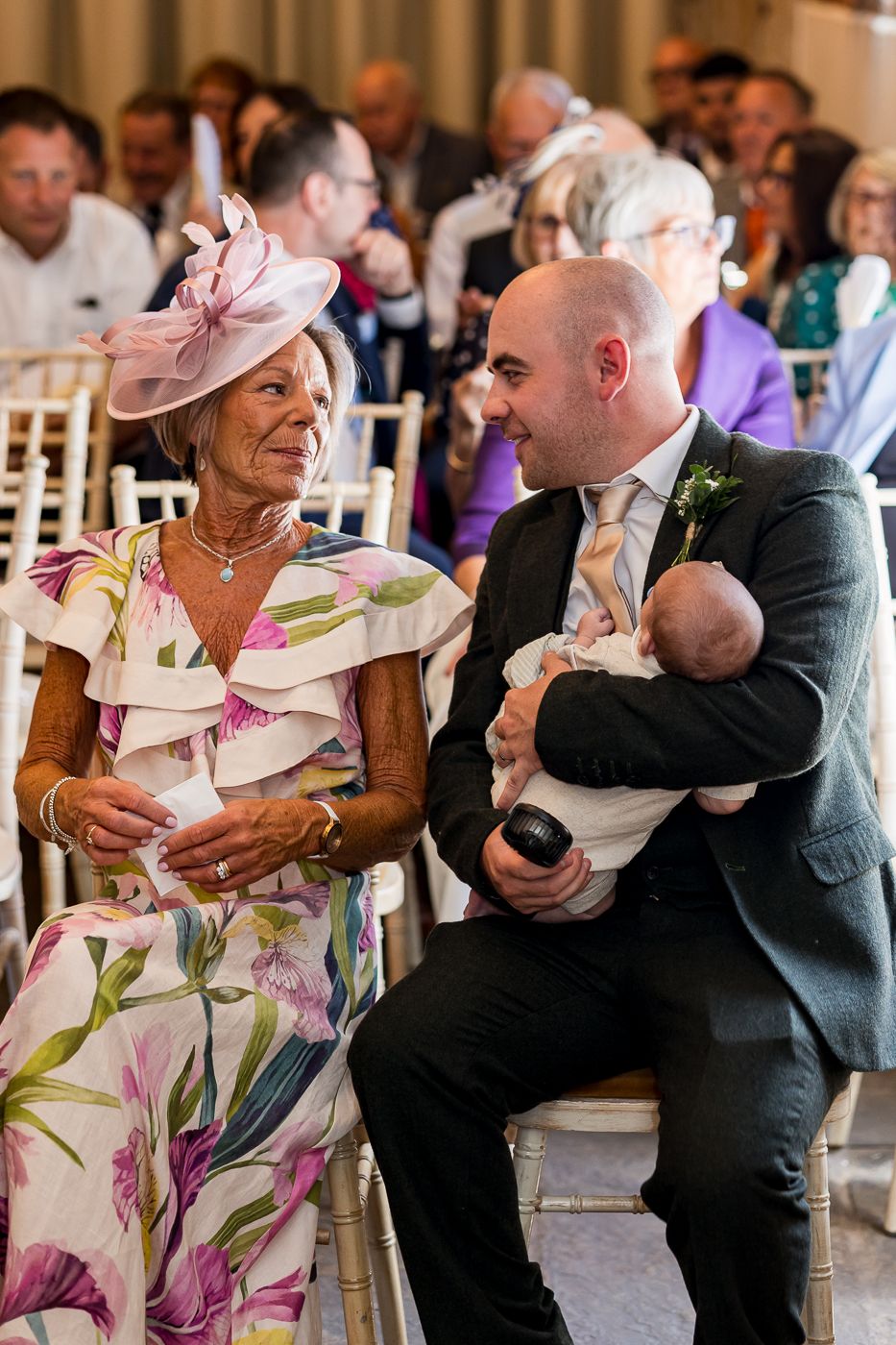 A man in a suit holds a baby while chatting with an elegantly dressed older woman in a floral outfit at a wedding.