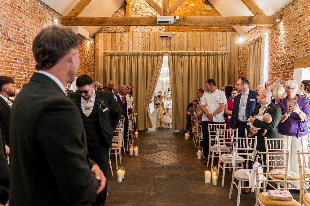 Elegant wedding ceremony in a rustic brick barn venue with candlelit aisle and guests seated on white chiavari chairs.