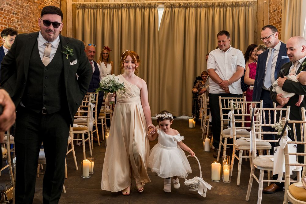 Bride walks down the aisle with flower girl at elegant indoor wedding ceremony with candles and white chairs.