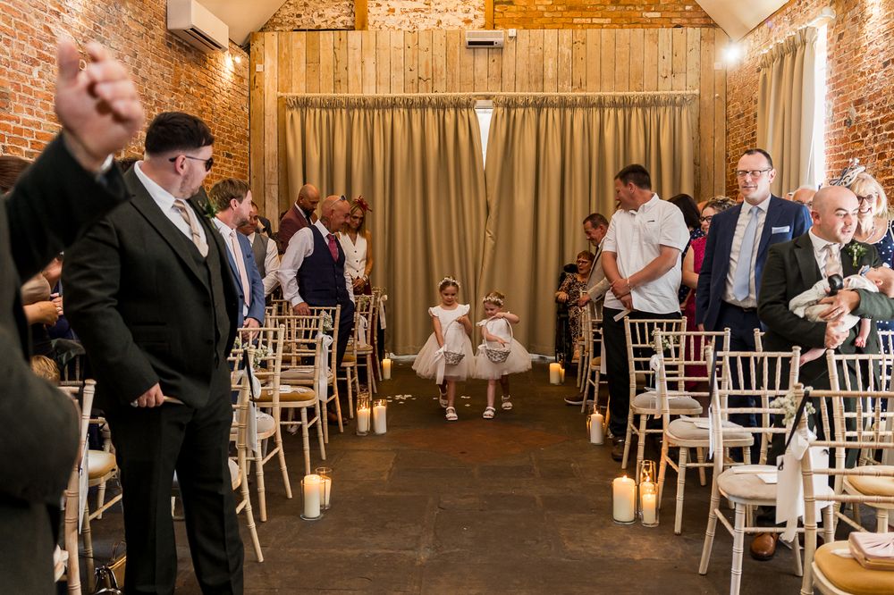 Flower girl walks down the aisle at an elegant barn wedding ceremony with candles and white chairs.