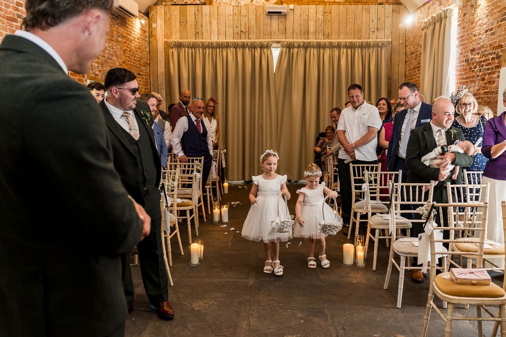 Two flower girls in white dresses walk down the aisle at an elegant rustic brick wedding venue.