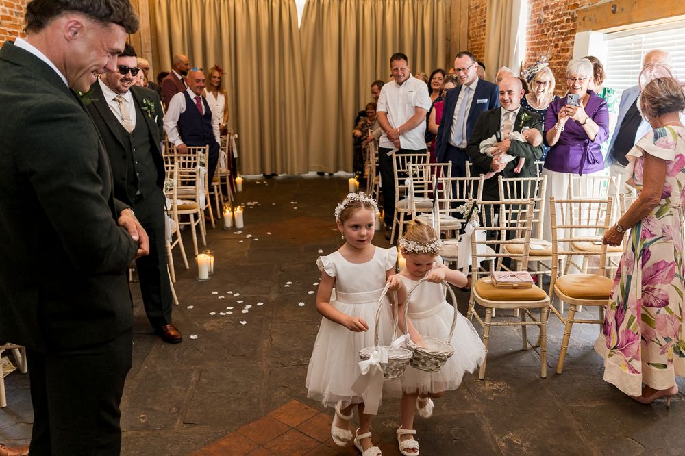 Flower girls scatter petals down the aisle as wedding guests applaud in a rustic barn venue.