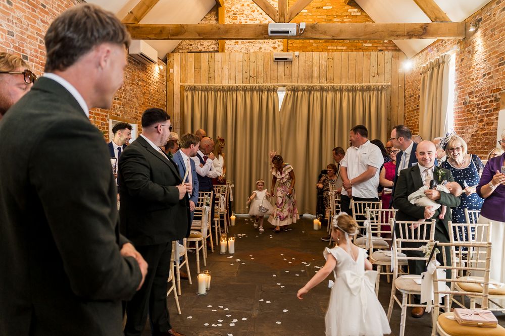 Flower girl scattering petals at an elegant barn wedding ceremony with guests seated on white chairs.