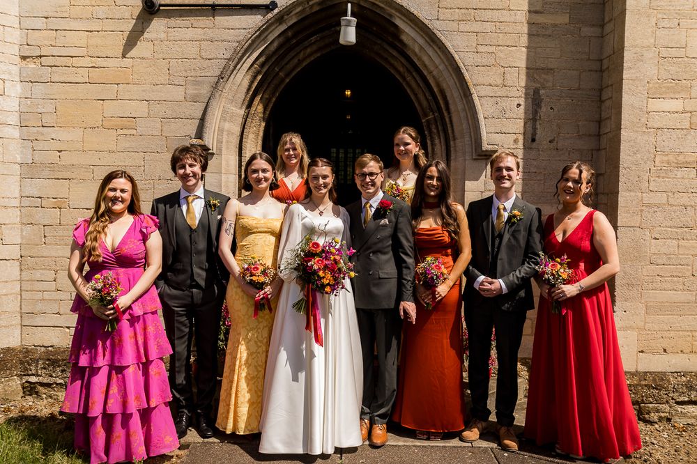 Wedding party posing outside stone church, bride in white gown with colorful bridesmaids in pink, gold, and red dresses.