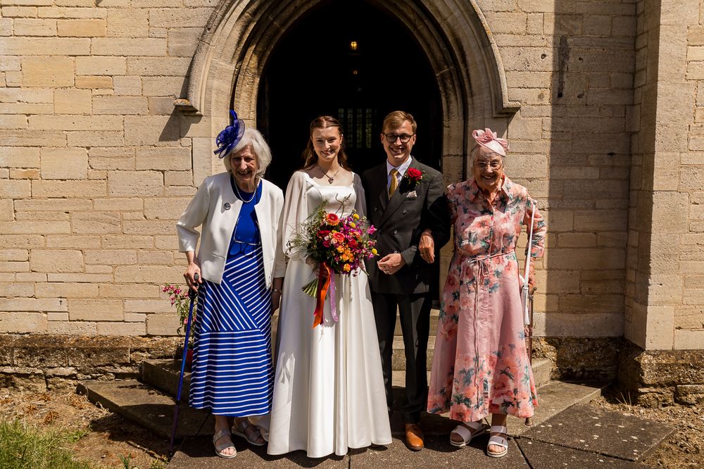 Bride and groom pose with two elderly guests outside a stone church on their sunny wedding day.