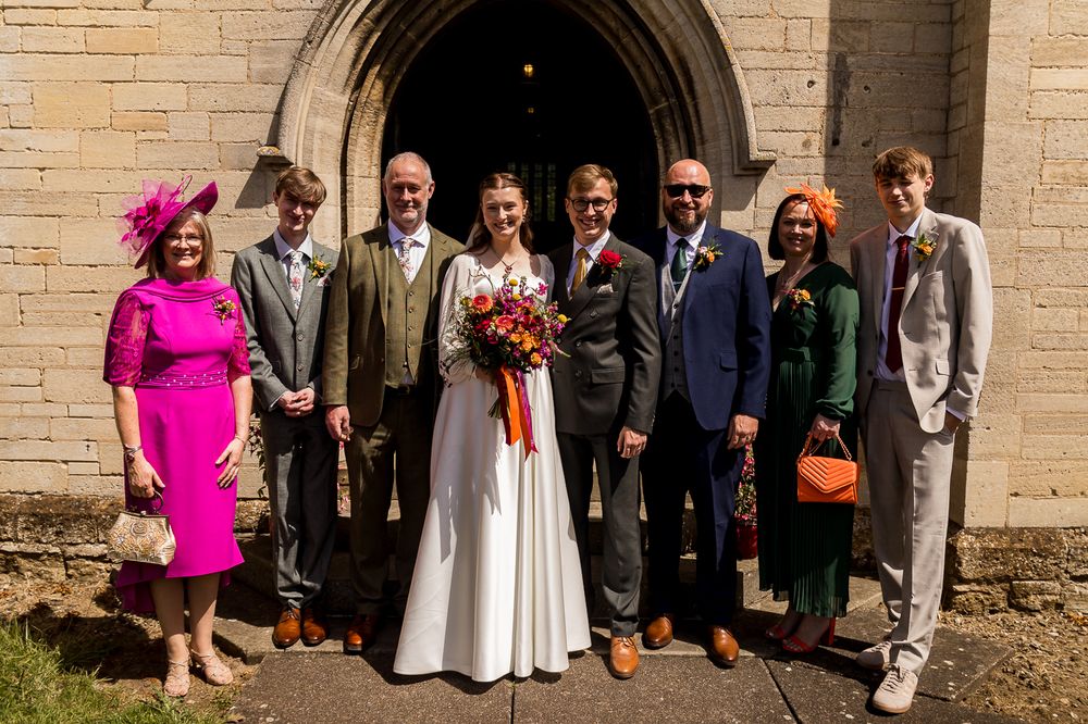 Wedding party posing outside a stone church, bride in white gown with groom and guests in colorful formal attire.
