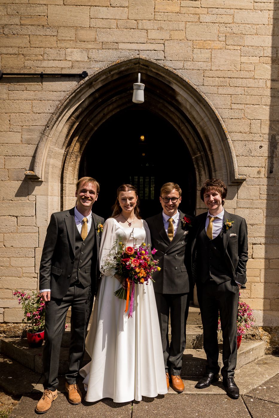 Bride in white gown with colorful bouquet poses with three groomsmen in dark suits outside a stone church.