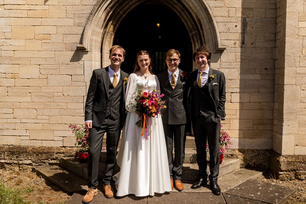 Bride and groom with two guests posing outside a stone church doorway, bride holding a colorful bouquet.