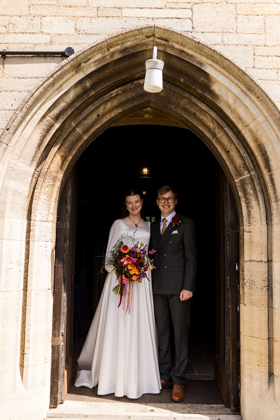 Bride and groom standing in a Gothic stone church doorway, bride holding a vibrant bouquet of colorful flowers.