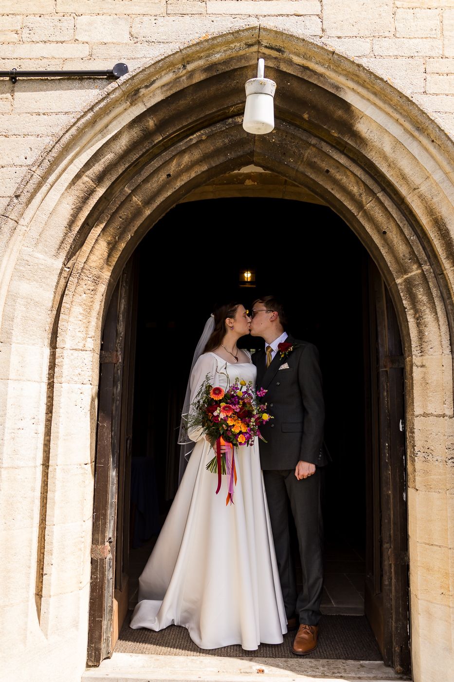 Bride and groom share a kiss in a stone church archway, bride holding a vibrant bouquet.