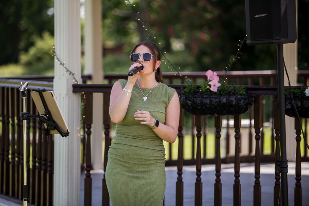 Woman in green dress singing into microphone outdoors at a gazebo event on a sunny day.