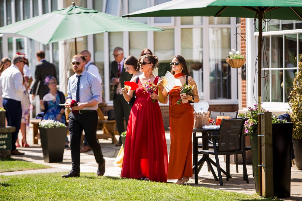 Bridesmaids in red and orange gowns holding drinks at an outdoor wedding reception on a sunny day.