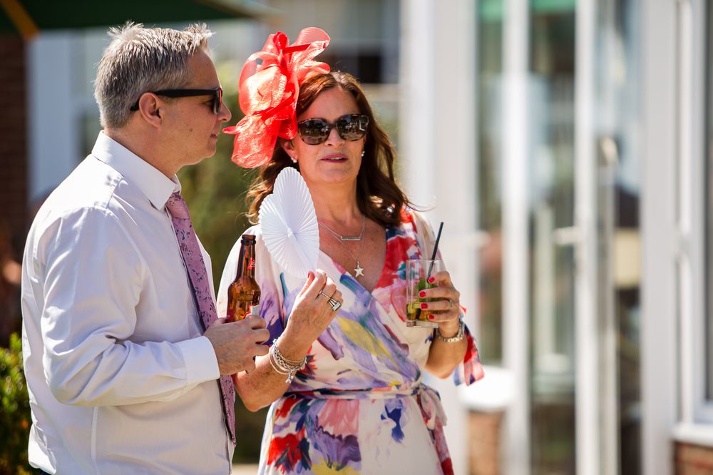 Elegantly dressed couple enjoying drinks outdoors, woman wearing floral dress and red fascinator hat at a summer event.
