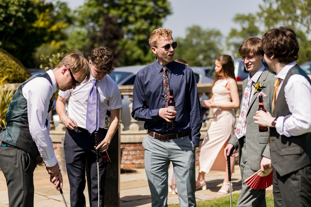Smartly dressed men socializing outdoors at a summer wedding celebration in a sunny garden setting.