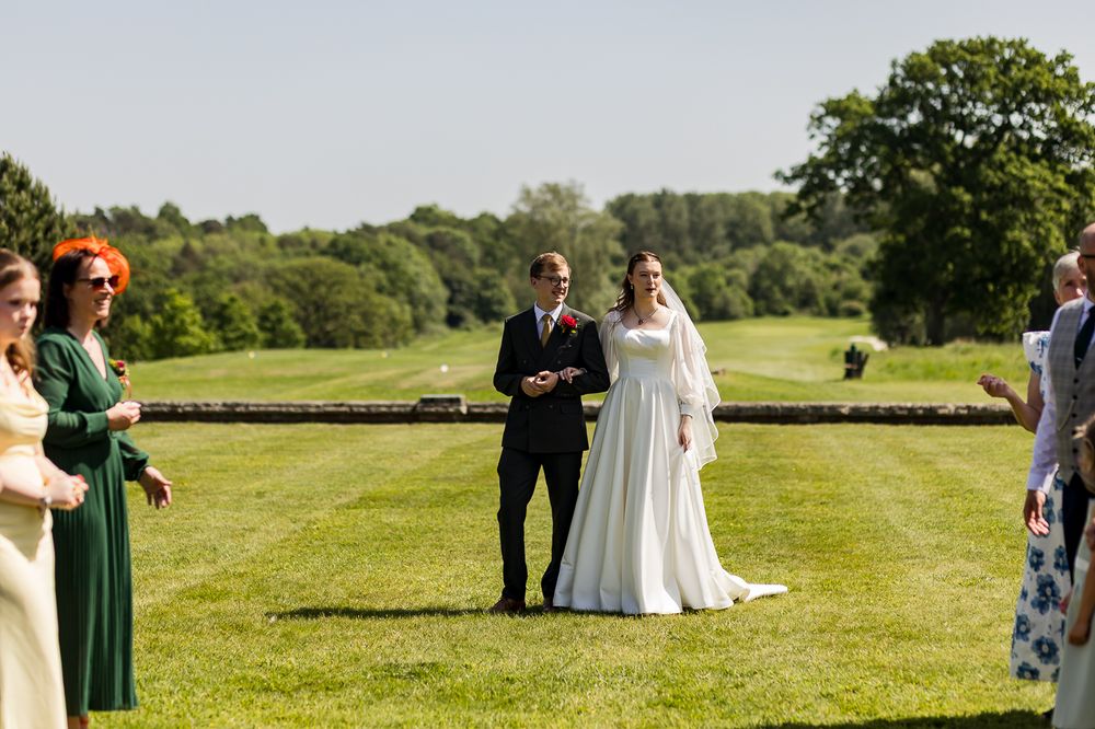 Bride and groom stand together during outdoor wedding ceremony on lush green lawn surrounded by guests on sunny day.