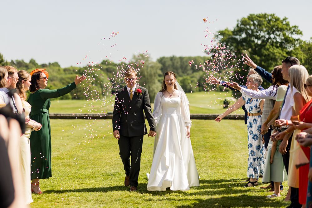 Bride and groom walk through confetti shower outdoors on sunny day, guests celebrating on both sides.