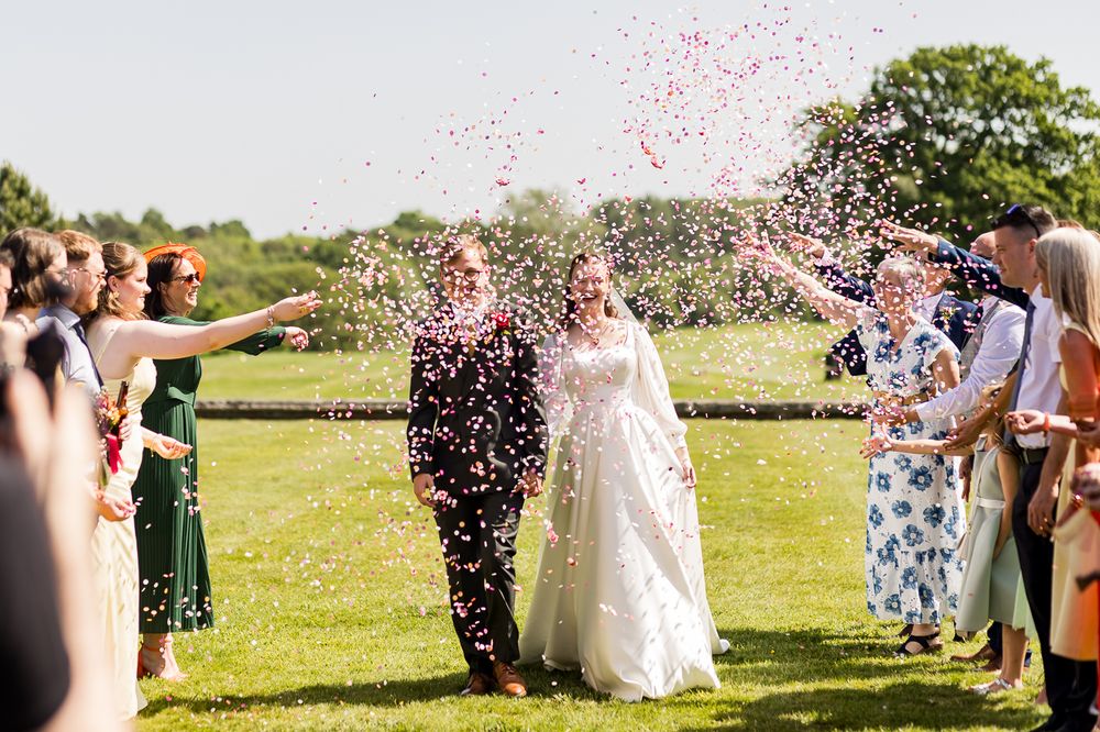 Newlyweds walk through a confetti shower surrounded by cheering guests on a sunny lawn.