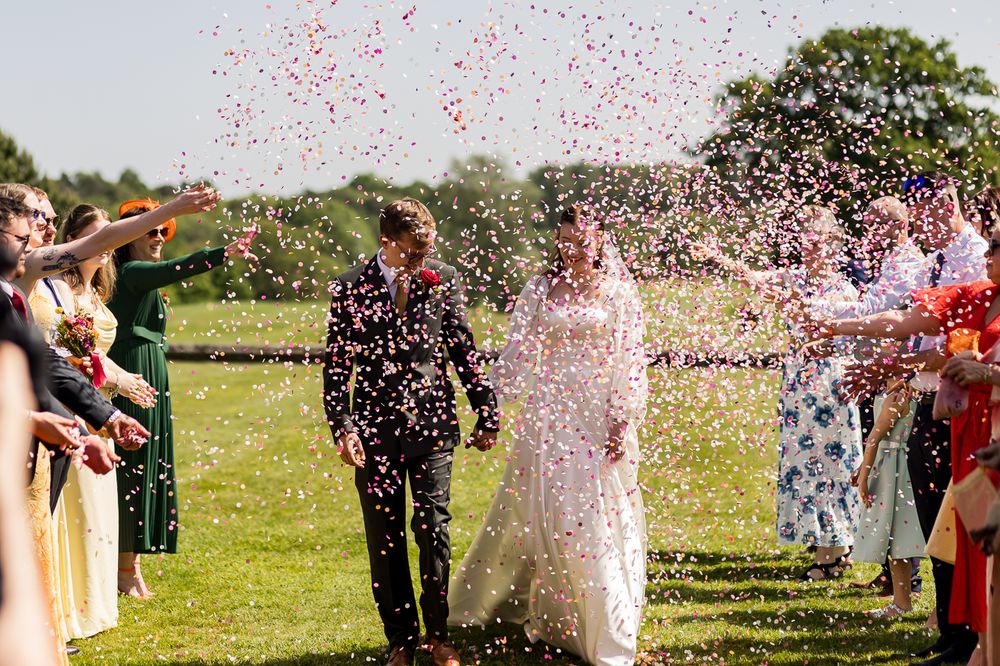 Bride and groom walk through confetti shower surrounded by cheering guests at outdoor summer wedding ceremony.