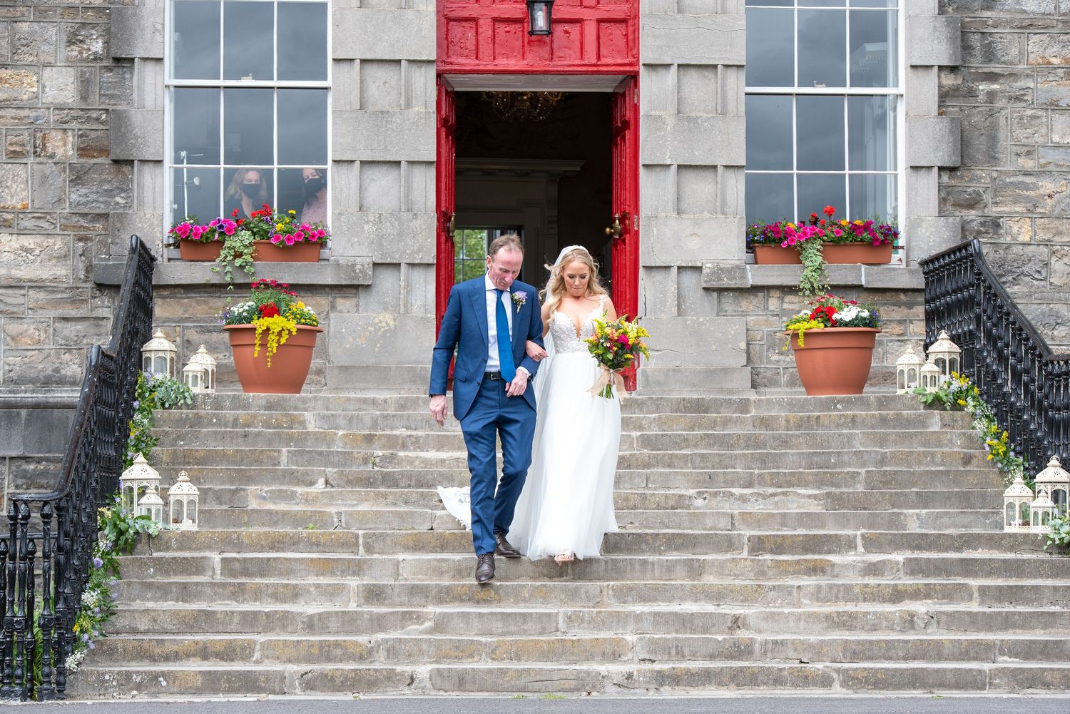Bride and groom descend stone steps of historic building with red door and floral arrangements on their wedding day at Bellinter house