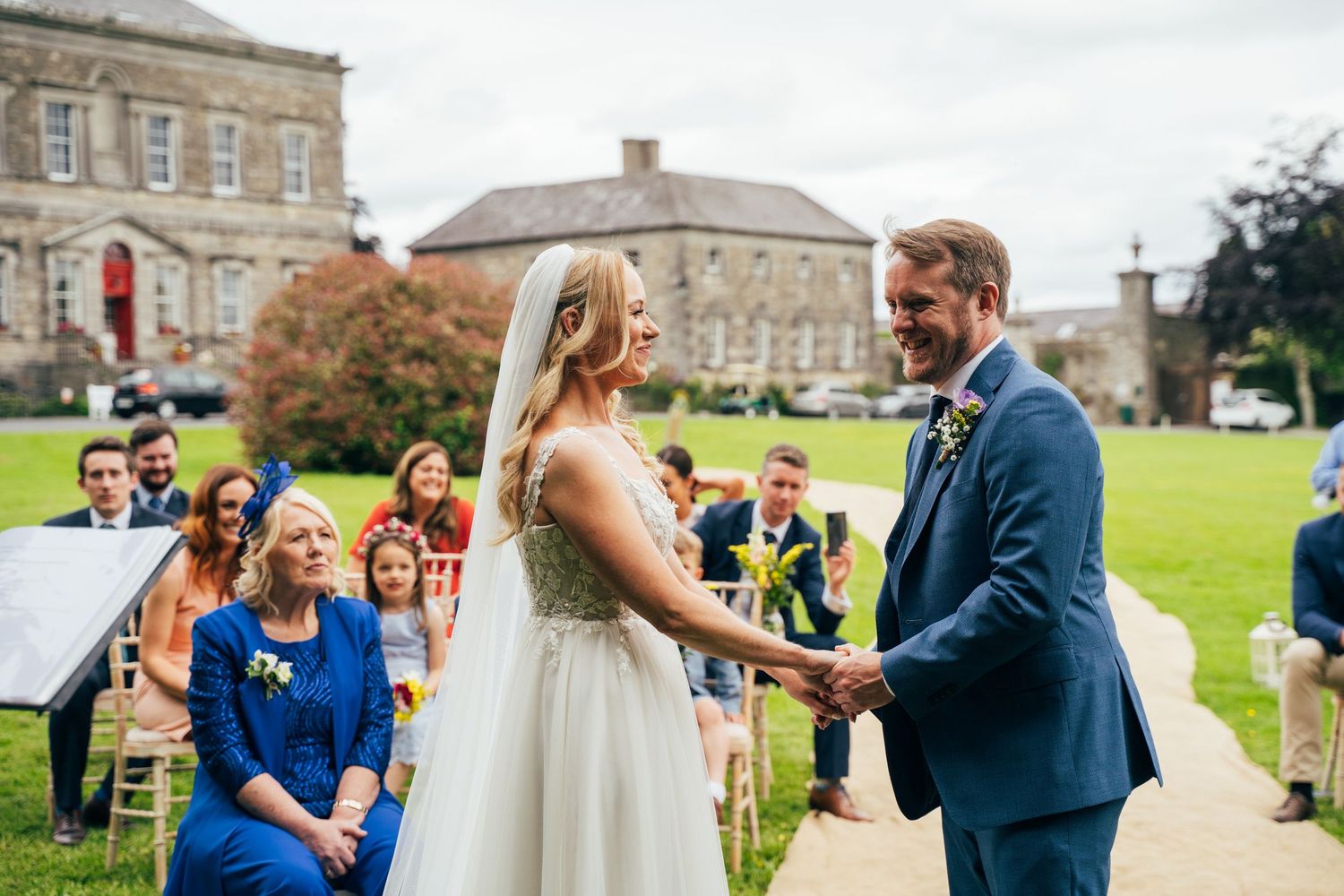 Bride and groom hold hands during outdoor wedding ceremony on garden path with guests seated beside historic Bellinter house