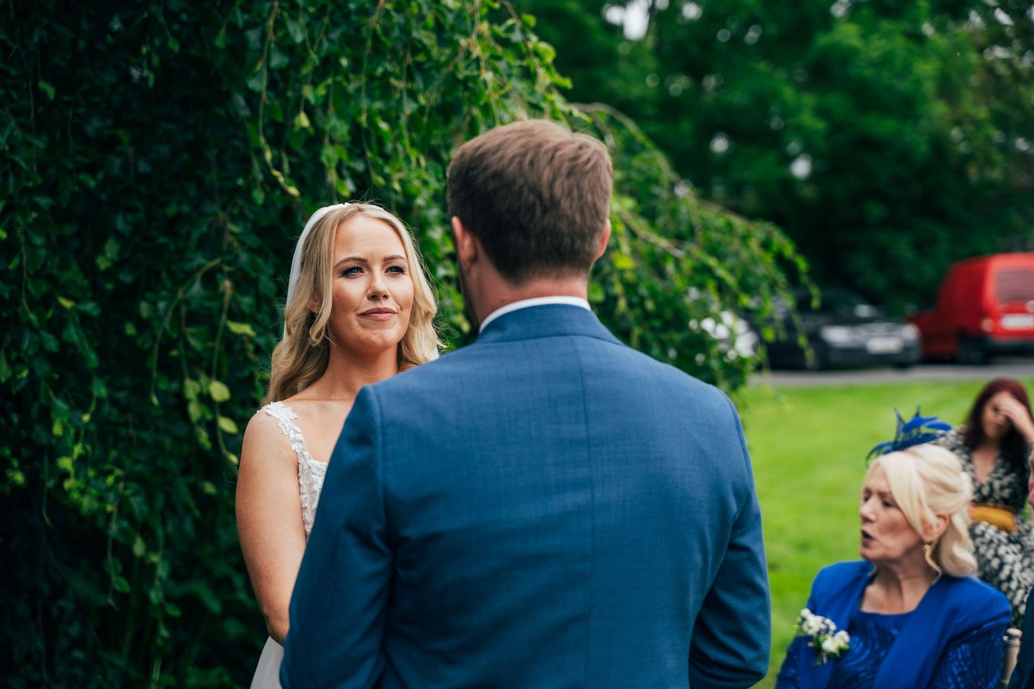 Bride and groom exchange vows outdoors during a wedding ceremony, with a guest in blue seated nearby at Bellinter house