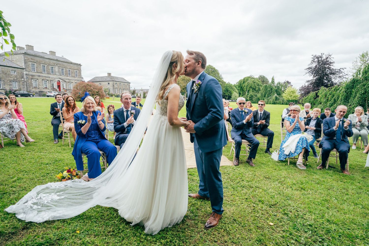 Bride and groom share first kiss at outdoor wedding ceremony on green lawn with guests applauding in background at historic Bellinter house.