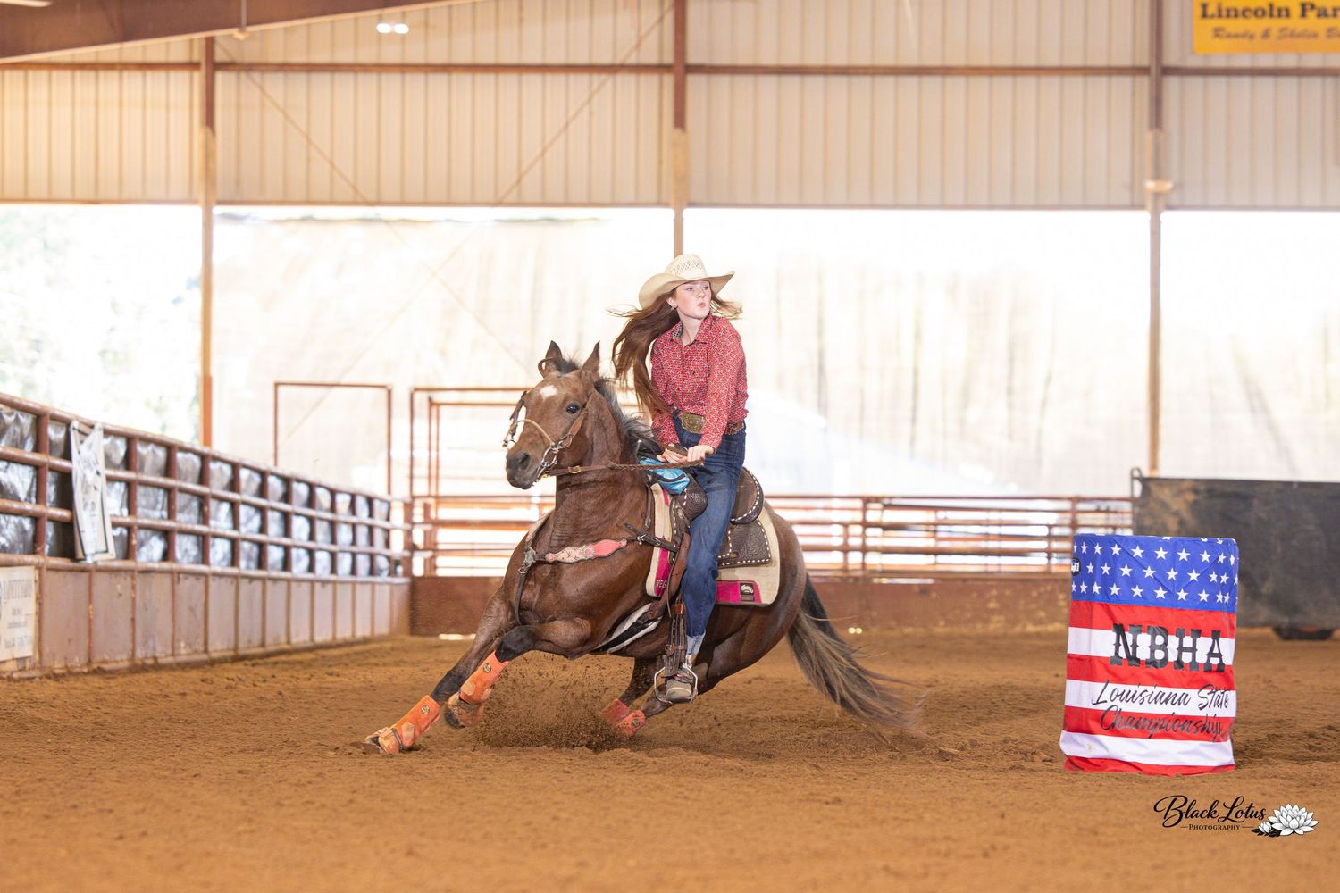 Barrel racer on a chestnut horse competing indoors at an NBHA rodeo event near a patriotic barrel.