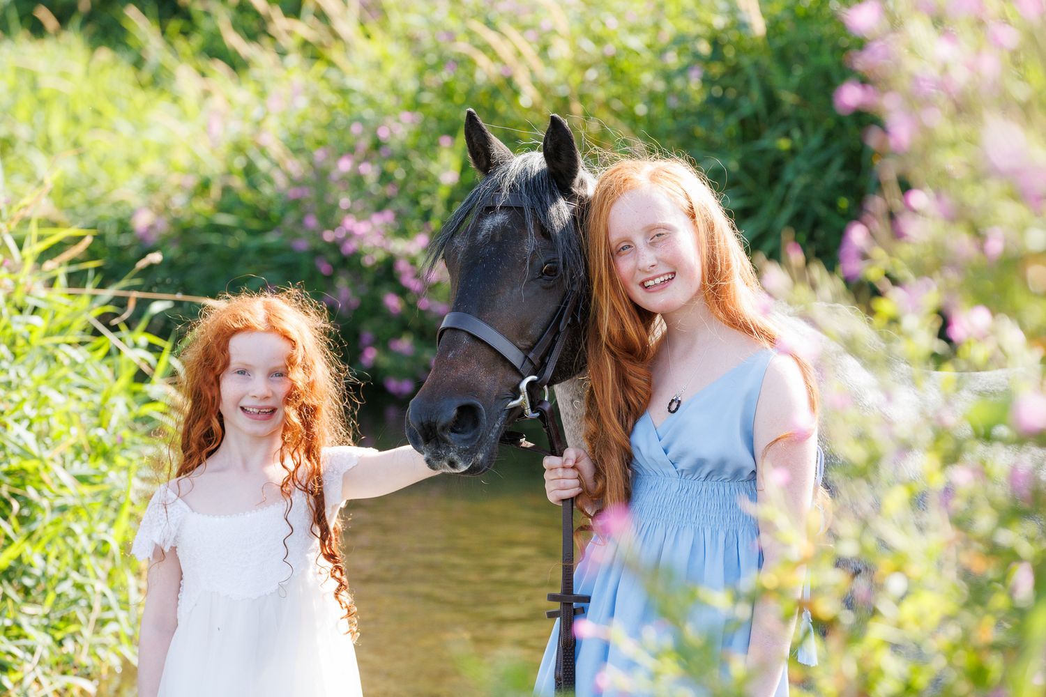 Young woman in black top smiling while gently embracing a brown horse outdoors among lush green trees.