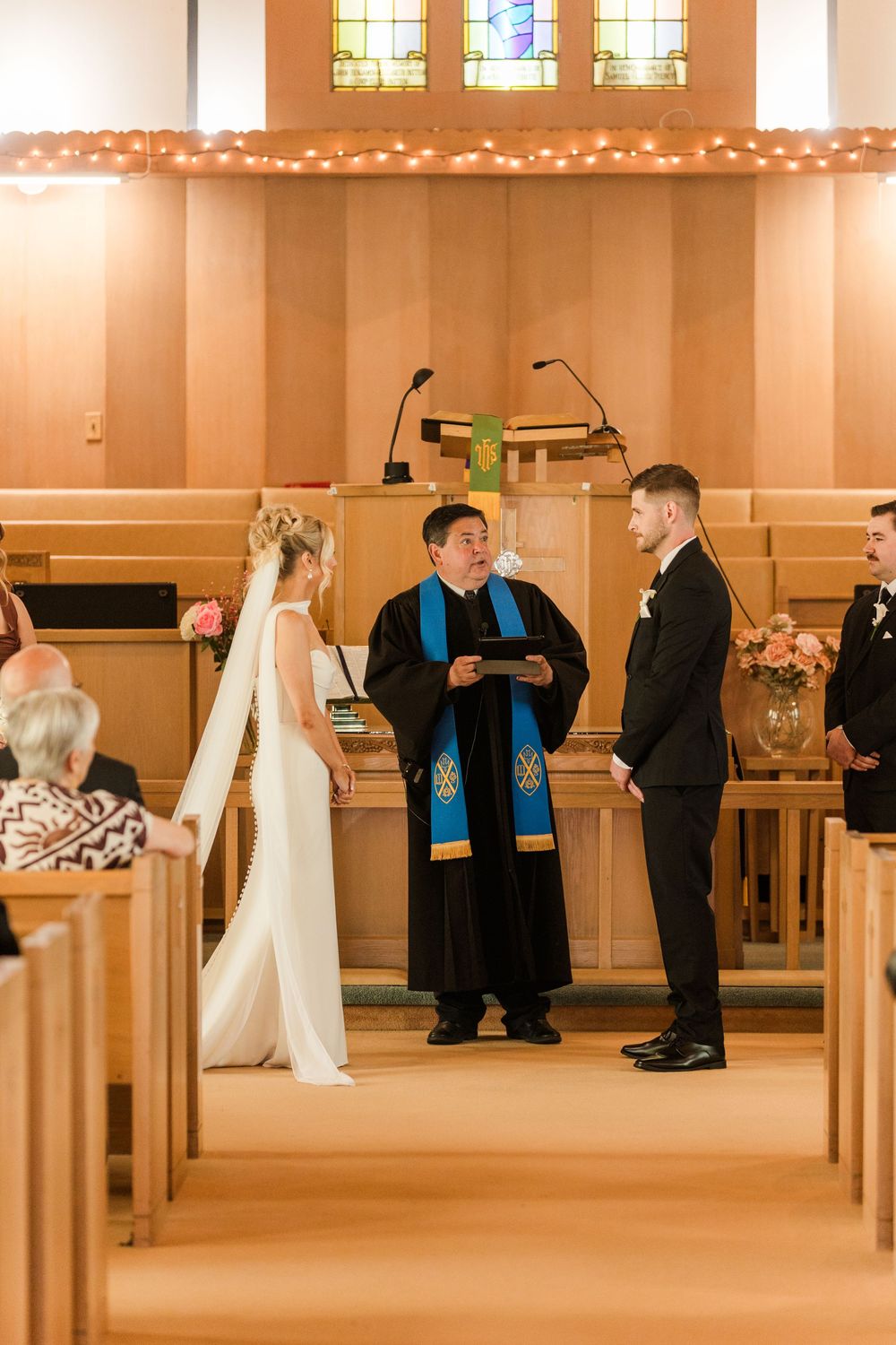 Bride and groom exchange vows before officiant in elegant wood-paneled church with stained glass windows and floral decor.