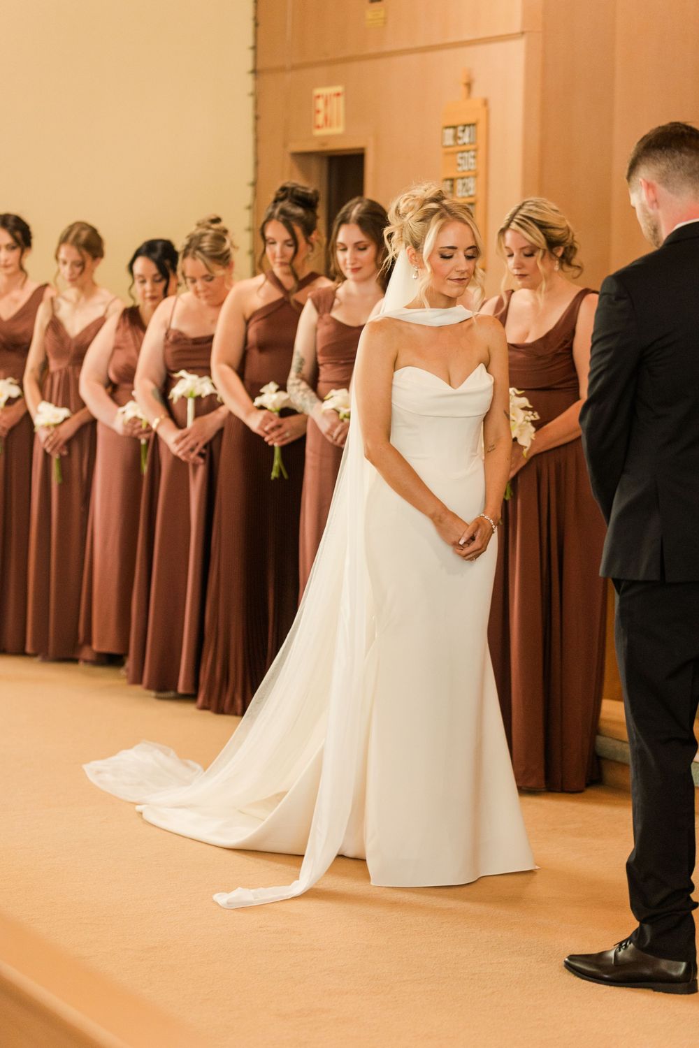 Bride in white gown stands with bridesmaids in rust dresses during church wedding ceremony prayer.