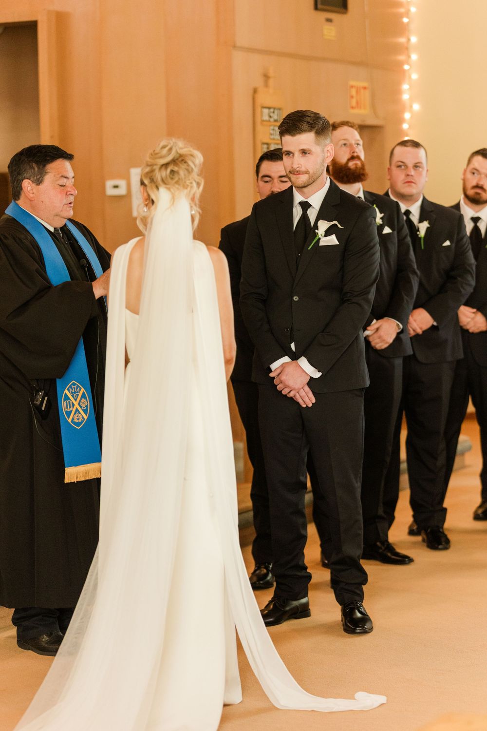 Groom in black suit smiles at bride in white cape wedding dress during church ceremony with groomsmen standing behind.