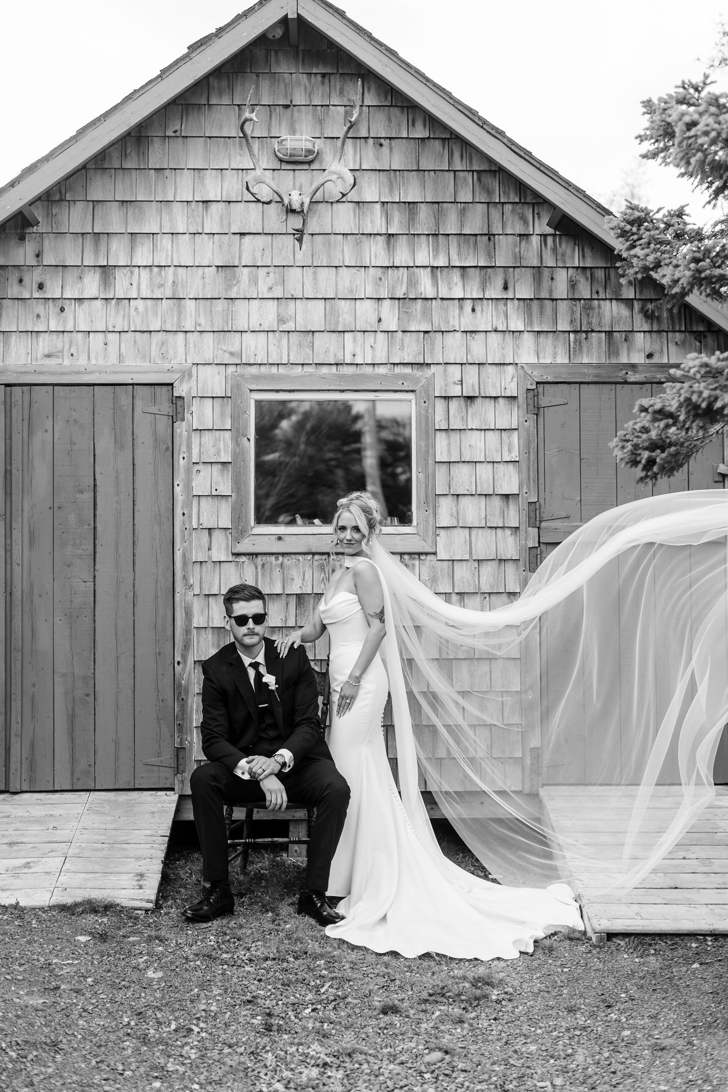 Black and white wedding photo of bride with flowing veil and groom in sunglasses seated outside rustic barn.