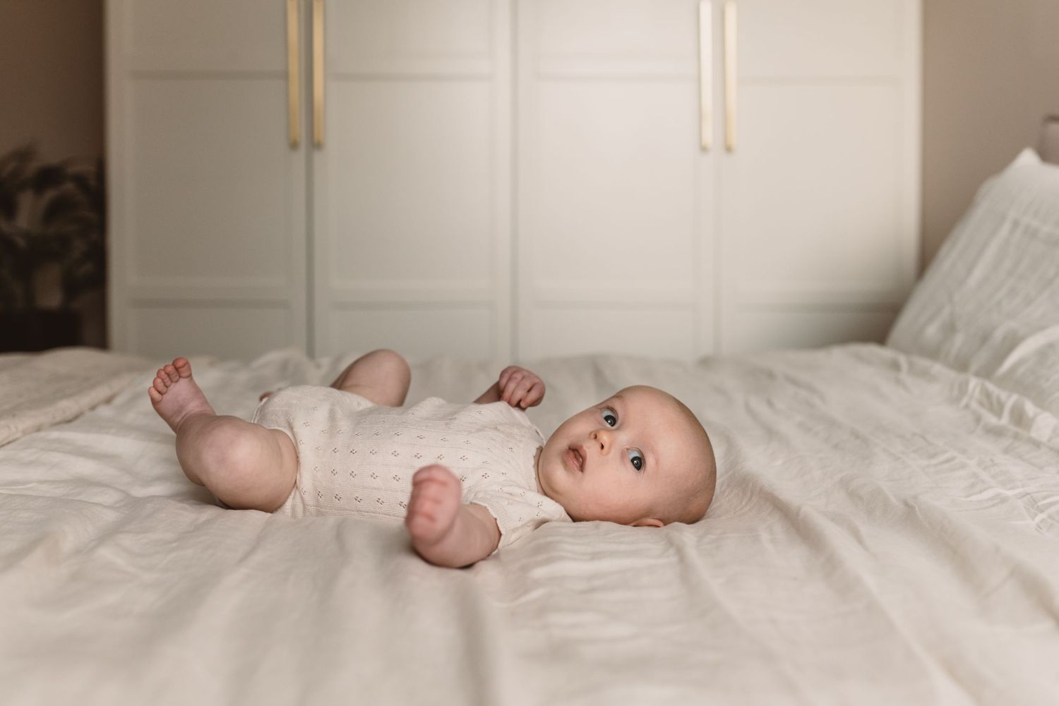 A cute baby in a white onesie lying on a bed with white bedding in front of a white wardrobe.