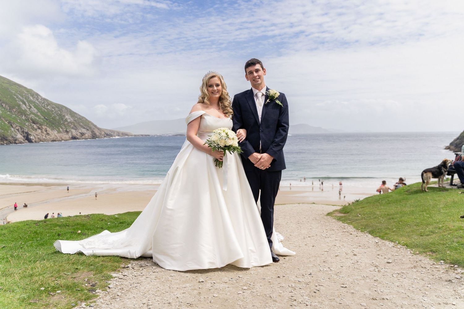 Bride and groom posing on a coastal path with a stunning beach and ocean backdrop on their wedding day.