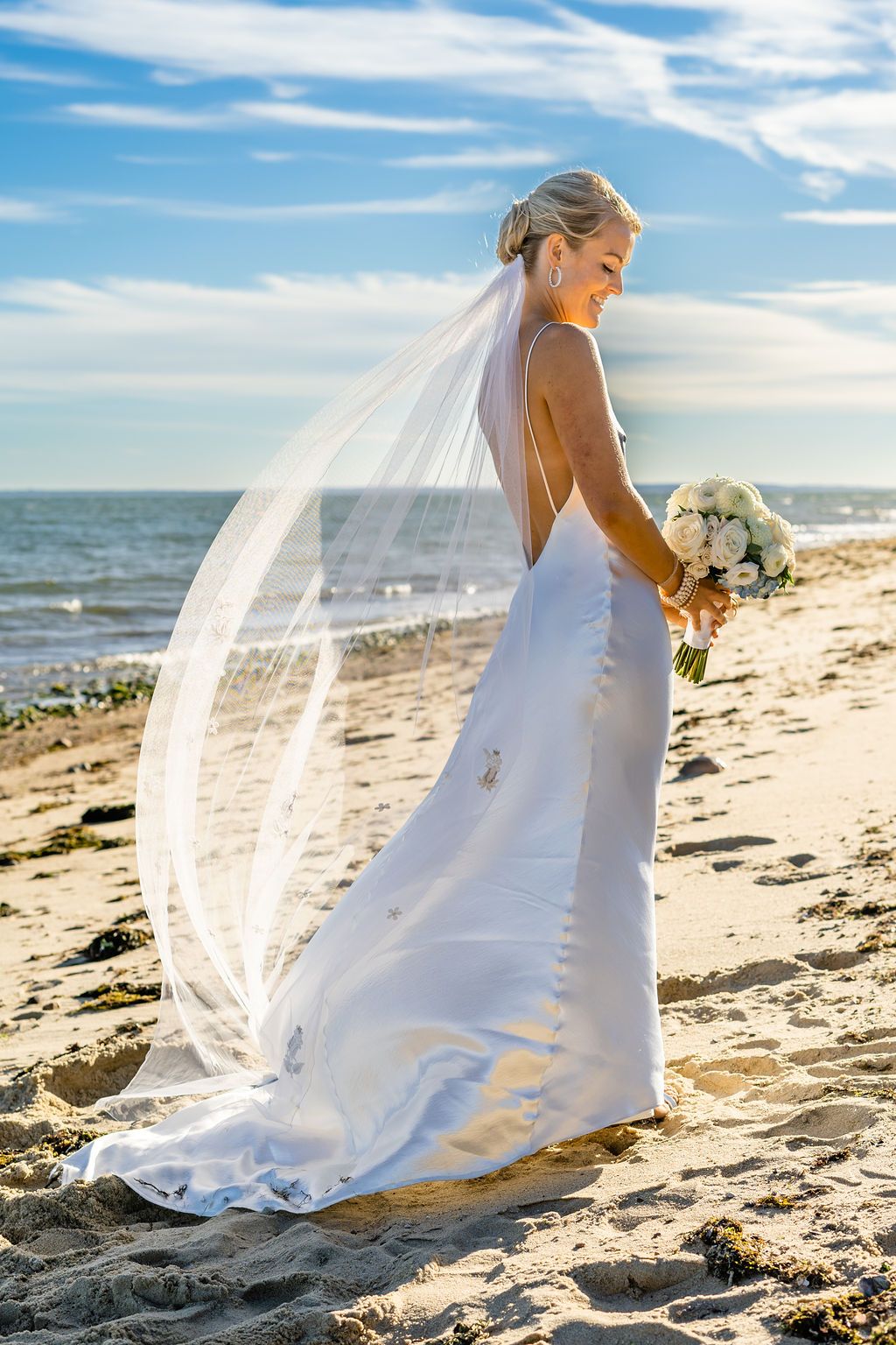 Bride in flowing white dress holding bouquet on beach with veil flowing in ocean breeze.