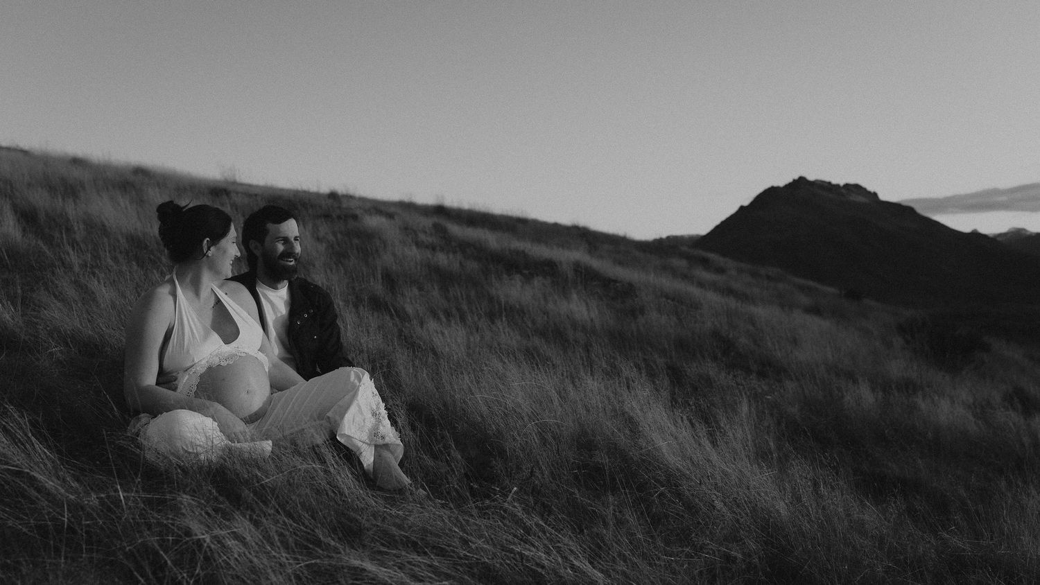 Expecting couple sits together on a grassy hillside at sunset in a black and white maternity photo with mountains behind them.