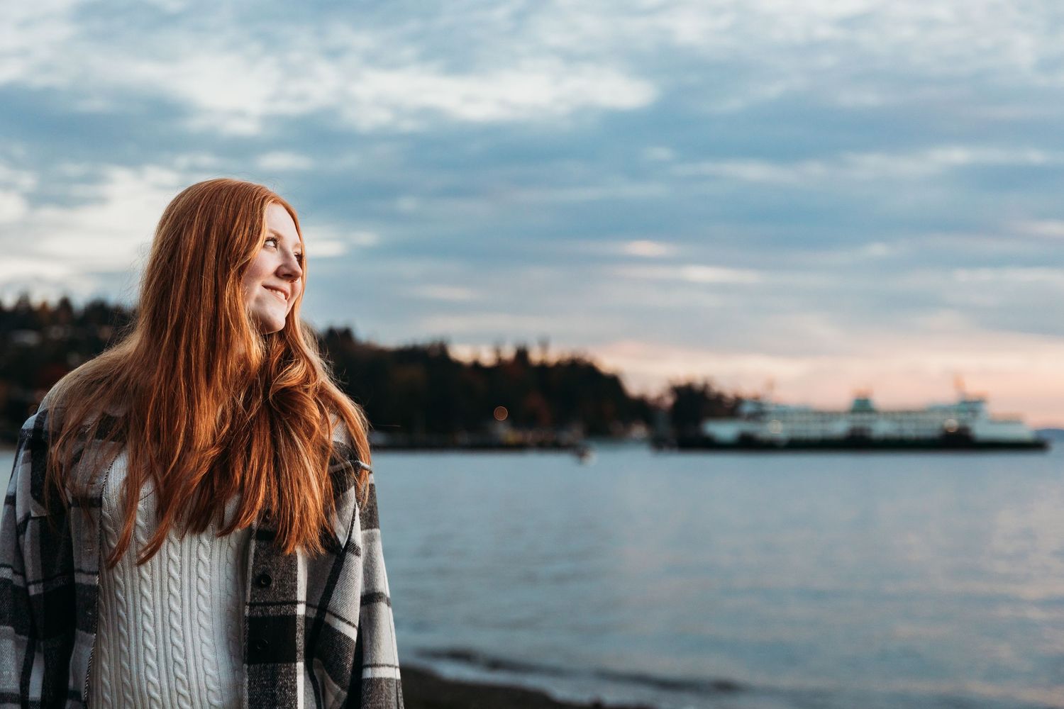 Smiling woman with long red hair in a plaid jacket gazes across calm water at sunset, with a ferry and forested shoreline in the distance.