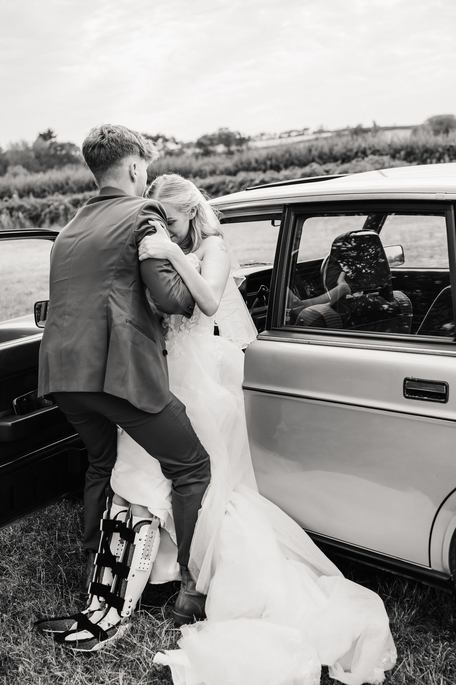 Black and white wedding photo of a bride in a flowing gown embracing a groom wearing a leg brace beside a vintage car in a grassy field.